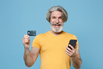 Cheerful elderly gray-haired mustache bearded man in casual yellow t-shirt posing isolated on blue background. People lifestyle concept. Mock up copy space. Hold credit bank card, using mobile phone.
