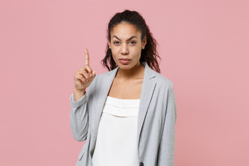 Displeased young african american business woman in grey suit white shirt posing isolated on pink wall background studio portrait. Achievement career wealth business concept. Pointing index finger up.