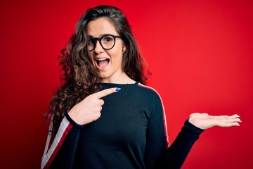 Young beautiful woman with curly hair wearing sweater and glasses over red background amazed and smiling to the camera while presenting with hand and pointing with finger.