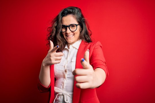 Young beautiful woman with curly hair wearing jacket and glasses over red background pointing fingers to camera with happy and funny face. Good energy and vibes.