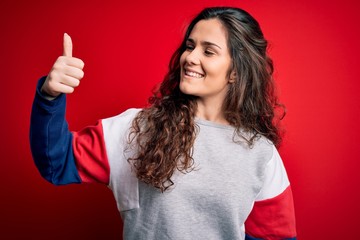 Young beautiful woman with curly hair wearing casual sweatshirt over isolated red background Looking proud, smiling doing thumbs up gesture to the side