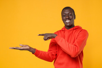 Smiling young african american man in red streetwear hoodie posing isolated on yellow wall background studio portrait. People lifestyle concept. Mock up copy space. Pointing index finger hand aside.
