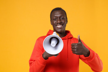 Smiling young african american man guy in red streetwear hoodie posing isolated on yellow background studio portrait. People lifestyle concept. Mock up copy space. Hold megaphone, showing thumb up.