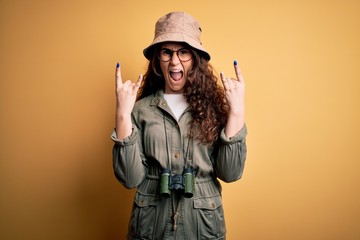 Young beautiful tourist woman on vacation wearing explorer hat and binoculars shouting with crazy expression doing rock symbol with hands up. Music star. Heavy concept.