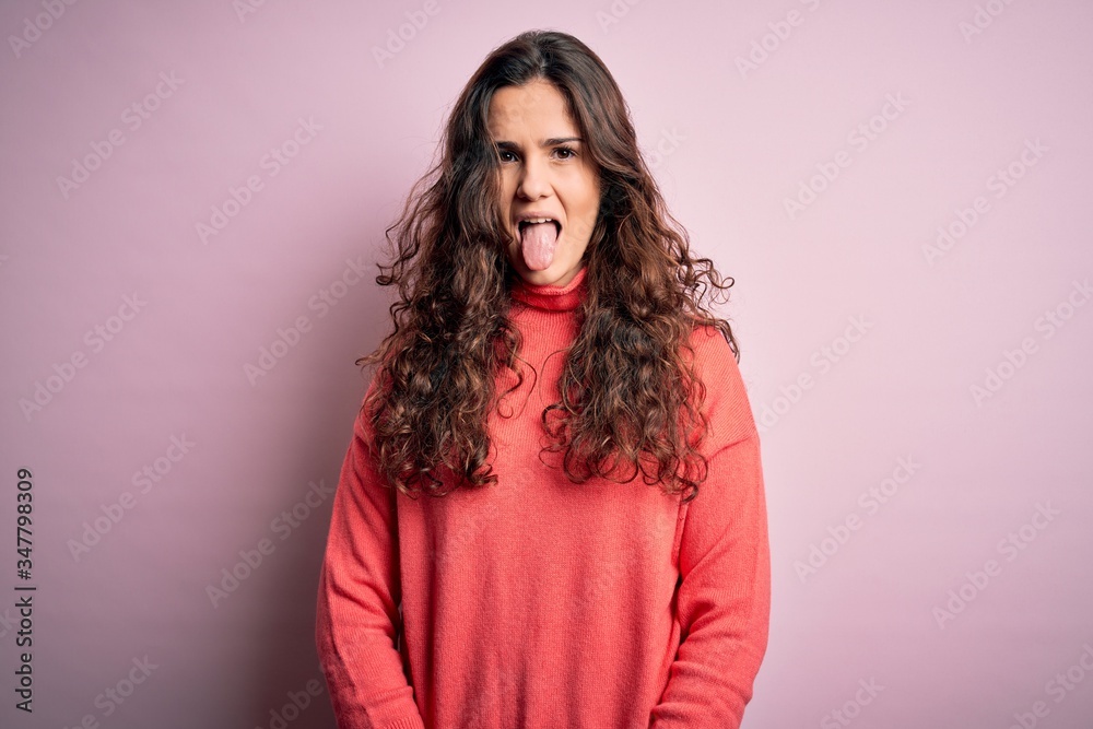 Poster Young beautiful woman with curly hair wearing turtleneck sweater over pink background sticking tongue out happy with funny expression. Emotion concept.