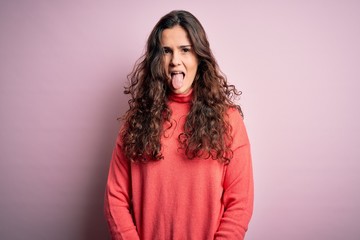 Fototapeta premium Young beautiful woman with curly hair wearing turtleneck sweater over pink background sticking tongue out happy with funny expression. Emotion concept.