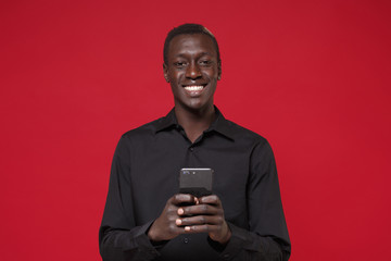 Smiling young african american man guy in classic black shirt posing isolated on red background studio portrait. People lifestyle concept. Mock up copy space. Using mobile phone, typing sms message.