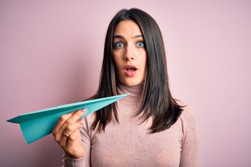 Young woman with blue eyes holding paper airplane standing over isolated pink background scared in shock with a surprise face, afraid and excited with fear expression