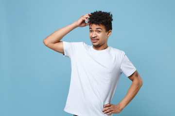 Preoccupied young african american guy in casual white t-shirt posing isolated on pastel blue background studio portrait. People lifestyle concept. Mock up copy space. Put hand on head, looking aside.