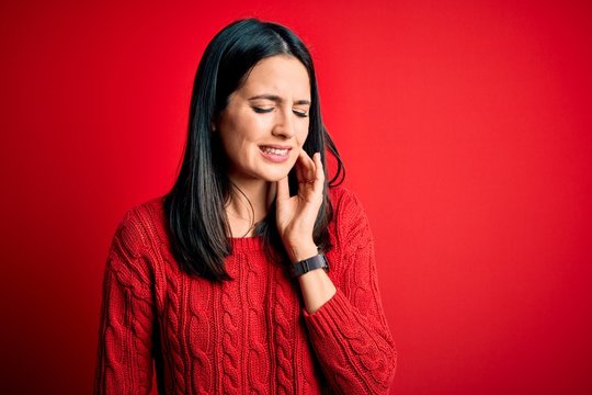 Young brunette woman with blue eyes wearing casual sweater over isolated red background touching mouth with hand with painful expression because of toothache or dental illness on teeth. Dentist