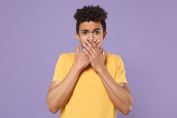 Fototapeta na wymiar Concerned young african american guy in casual yellow t-shirt posing isolated on pastel violet wall background studio portrait. People lifestyle concept. Mock up copy space. Covering mouth with hands.