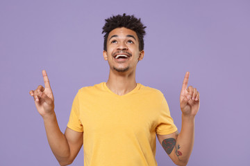 Cheerful young african american guy in casual yellow t-shirt posing isolated on pastel violet wall background studio portrait. People lifestyle concept. Mock up copy space. Pointing index fingers up.