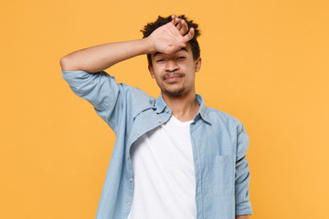 Exhausted young african american guy in casual blue shirt posing isolated on yellow wall background studio portrait. People sincere emotions lifestyle concept. Mock up copy space. Put hand on head.