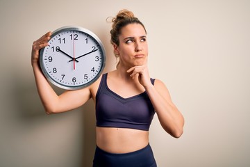 Young beautiful blonde sportswoman wearing sportswear on time to do sport holding clock serious face thinking about question, very confused idea