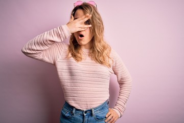 Young beautiful blonde woman wearing casual sweater and sunglasses over pink background peeking in shock covering face and eyes with hand, looking through fingers with embarrassed expression.