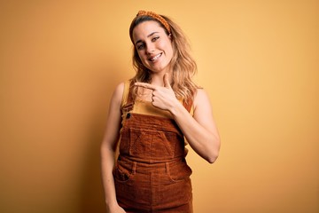 Young beautiful blonde woman wearing overalls and diadem standing over yellow background cheerful with a smile on face pointing with hand and finger up to the side with happy and natural expression