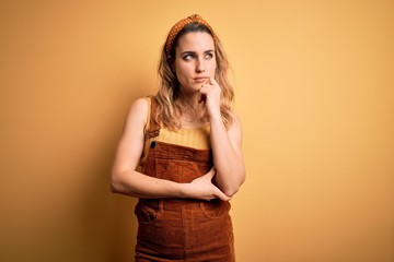 Young beautiful blonde woman wearing overalls and diadem standing over yellow background with hand on chin thinking about question, pensive expression. Smiling and thoughtful face. Doubt concept.