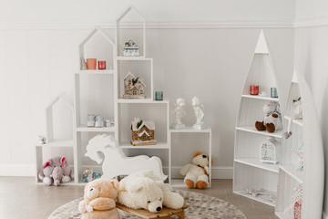 White Scandinavian style children's room interior with soft toys and wooden house-shaped shelves and white wooden rocking horse.