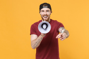 Laughing young bearded tattooed man guy in casual t-shirt black cap isolated on yellow background. People lifestyle concept. Mock up copy space. Screaming in megaphone pointing index finger on camera.
