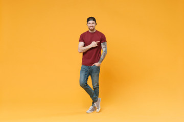 Fototapeta na wymiar Smiling young bearded tattooed man guy in casual t-shirt black cap posing isolated on yellow wall background studio portrait. People lifestyle concept. Mock up copy space. Pointing index finger aside.