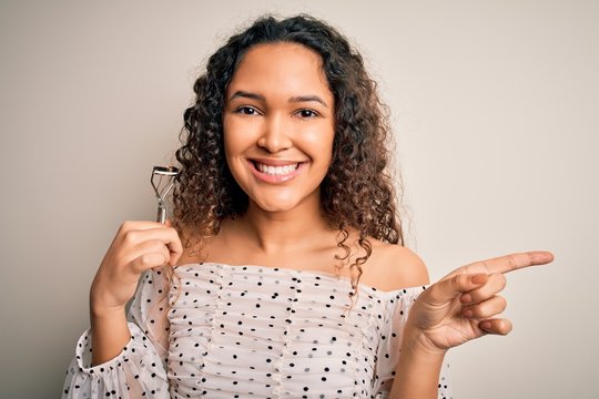 Young beautiful woman with curly hair holding eyelases curler over white background very happy pointing with hand and finger to the side