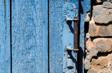 Old wooden door with a metal rusty handle. The surface of the door is painted blue, peeling off over time. The door is in the wall with sloppy red brick laid.