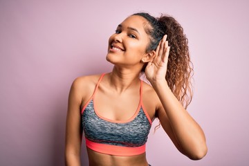 Fototapeta premium Beautiful sportswoman with curly hair doing sport wearing sportswear over pink background smiling with hand over ear listening an hearing to rumor or gossip. Deafness concept.