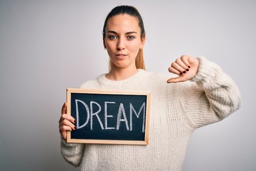 Young beautiful blonde woman with blue eyes holding blackboard with dream word message with angry face, negative sign showing dislike with thumbs down, rejection concept