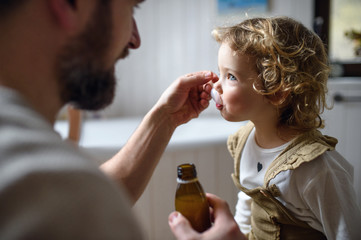 Unrecognizable father giving syrup to small sick daughter indoors at home.