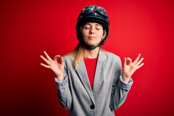 Young beautiful blonde motorcyclist woman wearing motorcycle helmet over red background relax and smiling with eyes closed doing meditation gesture with fingers. Yoga concept.