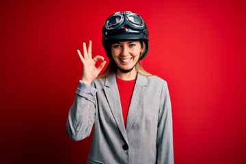Young beautiful blonde motorcyclist woman wearing motorcycle helmet over red background smiling positive doing ok sign with hand and fingers. Successful expression.