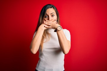 Beautiful blonde woman with blue eyes wearing casual white t-shirt over red background shocked covering mouth with hands for mistake. Secret concept.