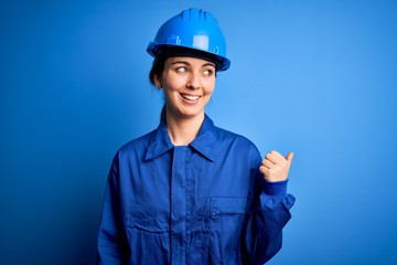 Young beautiful worker woman with blue eyes wearing security helmet and uniform smiling with happy face looking and pointing to the side with thumb up.