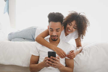 Young calm african loving couple girl guy in white t-shirts using mobile cell phone places his order online lying on bed indoors in bedroom at home, spend time in room. Quarantine lifestyle concept.