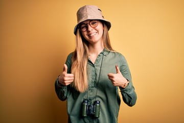 Beautiful blonde explorer woman with blue eyes wearing hat and glasses using binoculars success sign doing positive gesture with hand, thumbs up smiling and happy. Cheerful expression and winner