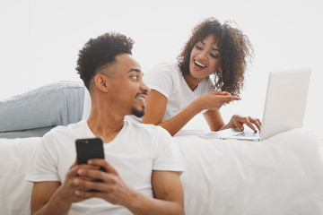 Young calm african loving couple girl guy in white t-shirts using mobile phone places his order online pc laptop lying on bed indoors in bedroom at home spend time in room Quarantine lifestyle concept