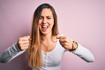 Young beautiful blonde woman with blue eyes wearing white t-shirt over pink background angry and mad raising fists frustrated and furious while shouting with anger. Rage and aggressive concept.