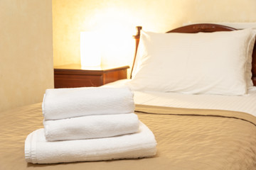 Two white towels are on the bed in hotel room. Stack of towels on a hotel bed is close-up, horizontal background. In the background a bed and a luminous lamp. Space for text