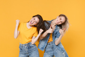 Two cheerful young women girls friends in casual t-shirts denim clothes isolated on yellow wall background studio portrait. People lifestyle concept. Clenching fists like winner, showing thumbs up.