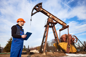 Petroleum engineer in work overalls and helmet holding clipboard, checking oil pumping unit, making notes. Worker standing near oil pump jack. Concept of oil extraction and petroleum industry.
