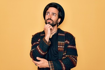 Young handsome hispanic bohemian man wearing hippie style and boho hat with hand on chin thinking about question, pensive expression. Smiling and thoughtful face. Doubt concept.