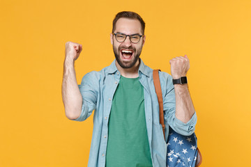 Joyful young man student in casual clothes glasses backpack posing isolated on yellow background. Education in high school university college concept. Wearing smart watch on hand doing winner gesture.