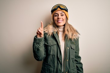 Young brunette skier woman wearing snow clothes and ski goggles over white background pointing finger up with successful idea. Exited and happy. Number one.