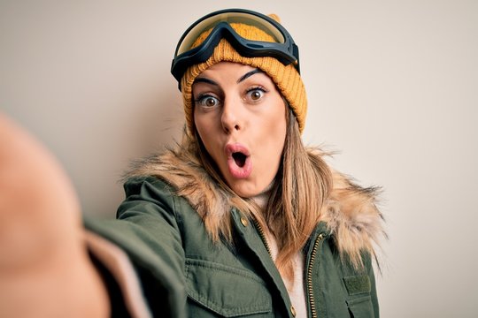 Young brunette skier woman wearing snow clothes and ski goggles make selfie by camera scared in shock with a surprise face, afraid and excited with fear expression