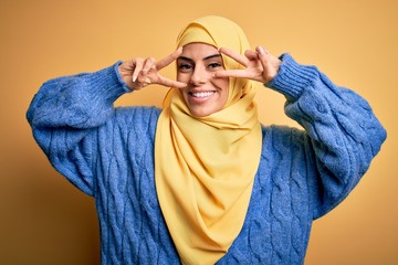 Young beautiful brunette muslim woman wearing arab hijab over isolated yellow background Doing peace symbol with fingers over face, smiling cheerful showing victory