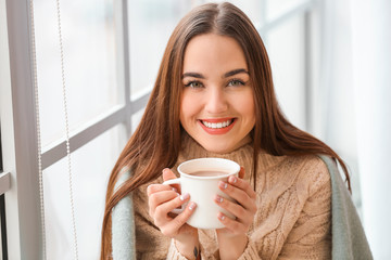 Beautiful young woman drinking hot cocoa near window at home