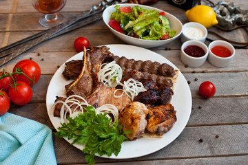 Assorted grilled meat on a white plate with herbs. Lots of meat on a wooden background with sauces and vegetables