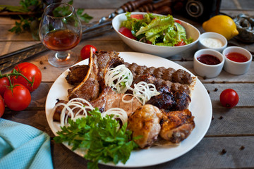Assorted grilled meat on a white plate with herbs. Lots of meat on a wooden background with sauces and vegetables