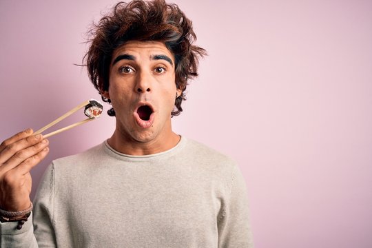 Young Handsome Man Eating Sushi Using Chopsticks Over Isolated Pink Background Scared In Shock With A Surprise Face, Afraid And Excited With Fear Expression