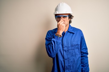 Young constructor man wearing uniform and security helmet over isolated white background smelling something stinky and disgusting, intolerable smell, holding breath with fingers on nose. Bad smell